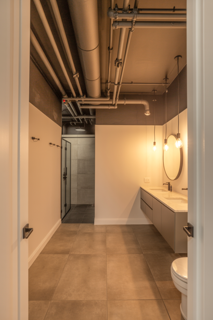 A basement bathroom with an industrial style matte black painted ceiling and exposed pipes contrasting against white walls, concrete-look floor tiles, and black matte fixtures throughout