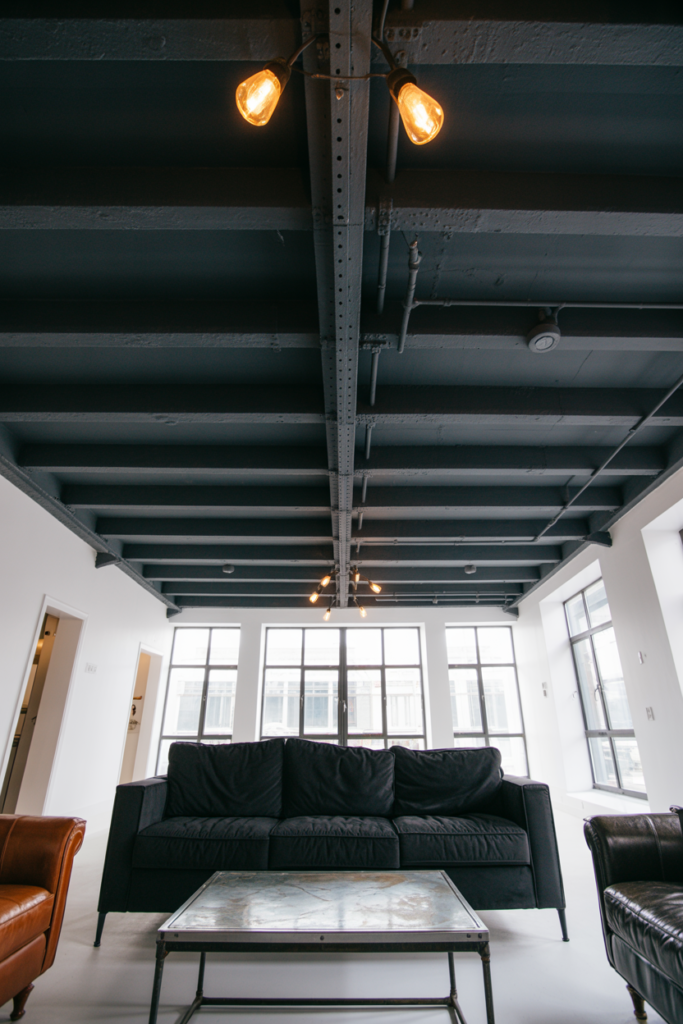 Exposed ceiling joists and ceiling void painted deep matte black in a high-ceilinged industrial living room with white walls and Edison pendant lights